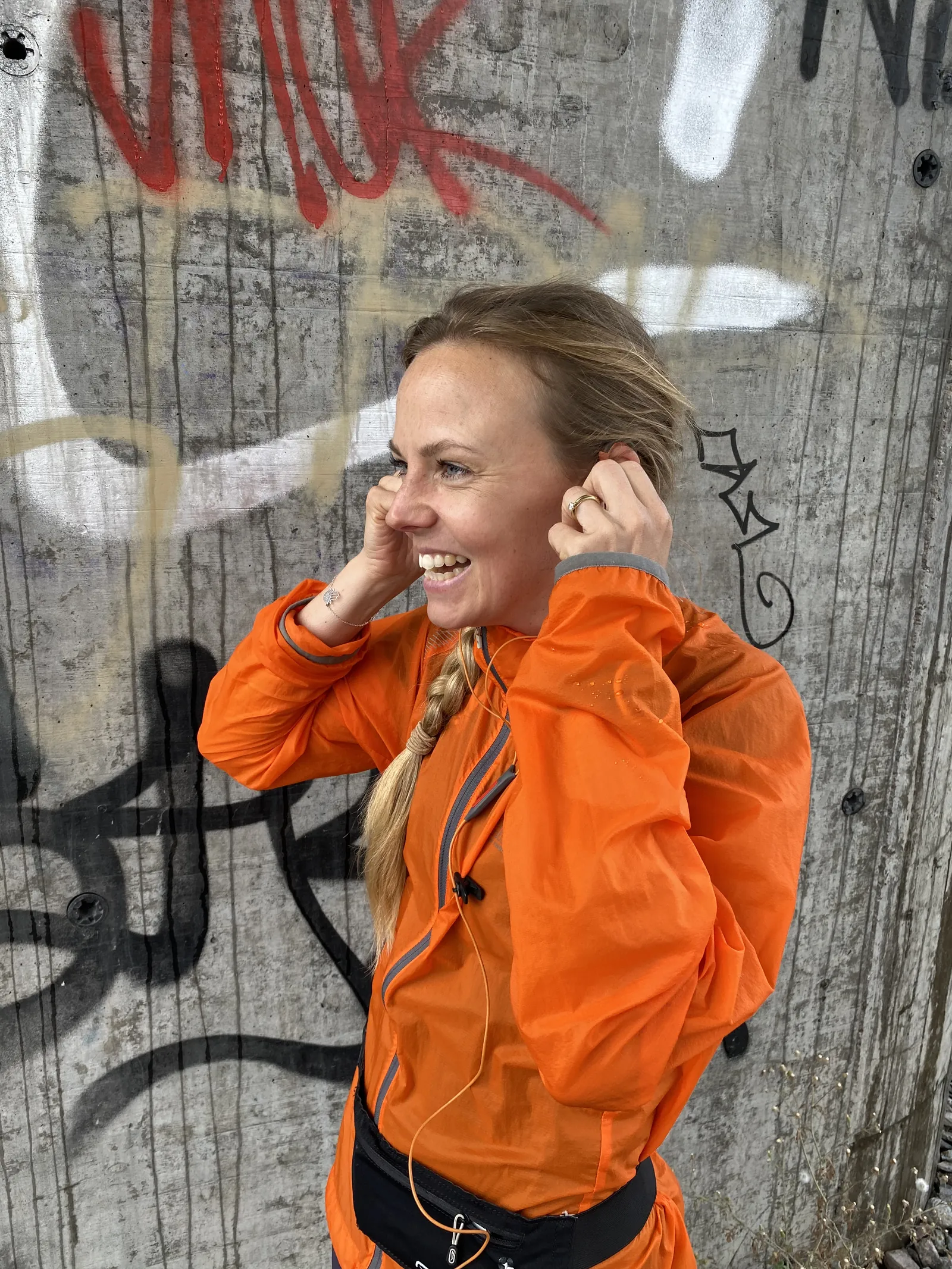 En löpare i orange jacka sätter i hörluren framför en graffitivägg